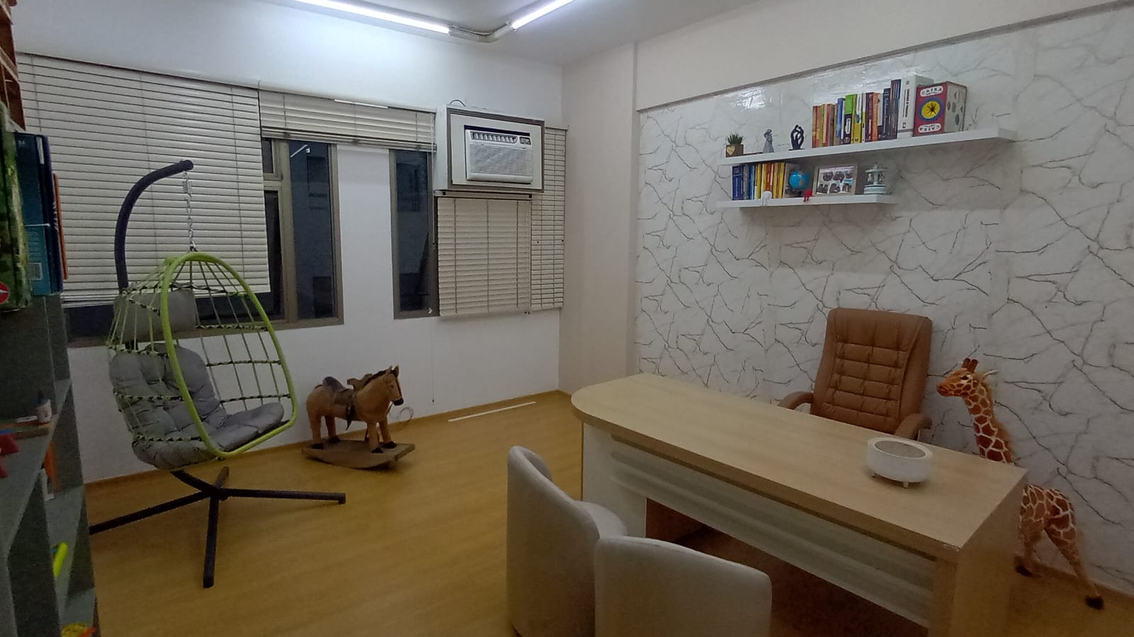 Consultório da Dra. Gisela Alves no Edifício Asteca, com poltrona suspensa, cavalinho de balanço, girafa de pelúcia, mesa de atendimento e estante com livros